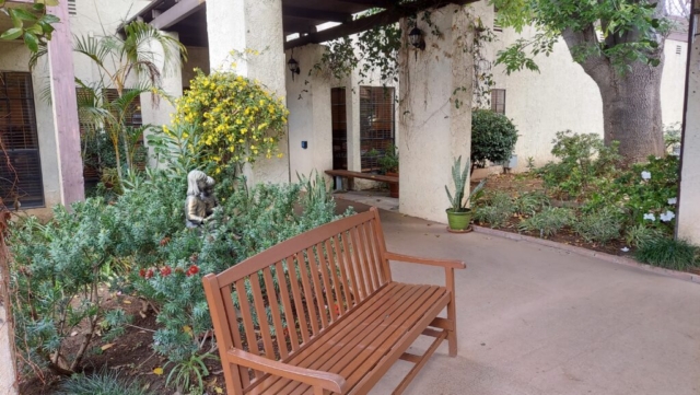 The front entrance to The Oaks of Pasadena features a wooden bench, statue and colorful garden